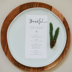 May include: A white card with black text on a white plate with a wooden charger. The card reads "thankful" and lists the menu for a Thanksgiving dinner. The menu includes roasted butternut squash soup, classic roast turkey, sausage and herb stuffing, baked mashed potatoes, honey glazed carrots, oven roasted broccoli, old fashioned pumpkin pie, pecan pie, and pumpkin chocolate chip cookies.