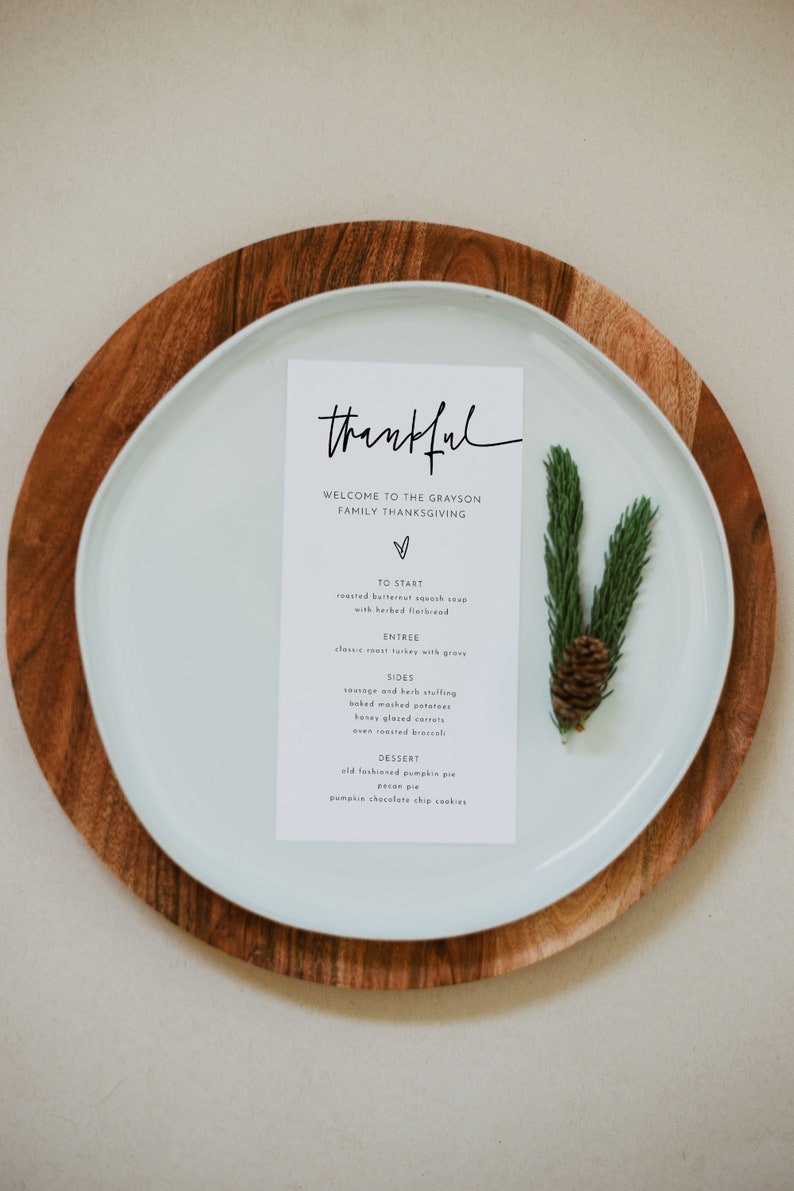 May include: A white card with black text on a white plate with a wooden charger. The card reads "thankful" and lists the menu for a Thanksgiving dinner. The menu includes roasted butternut squash soup, classic roast turkey, sausage and herb stuffing, baked mashed potatoes, honey glazed carrots, oven roasted broccoli, old fashioned pumpkin pie, pecan pie, and pumpkin chocolate chip cookies.