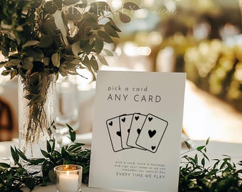 May include: A white sign with the text "pick a card ANY CARD" and a drawing of four playing cards with hearts. The sign is on a table with a candle and greenery. The text below reads "pick a card, write a message, and sign your name so we can remember our special day EVERY TIME WE PLAY."