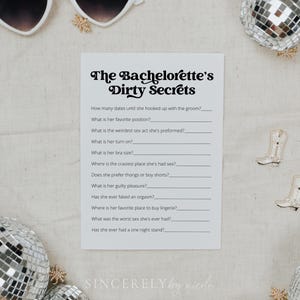 A printed game card for "The Bachelorette’s Dirty Secrets" resting on soft beige fabric with dried floral elements. A fun party activity designed for playful and candid moments.