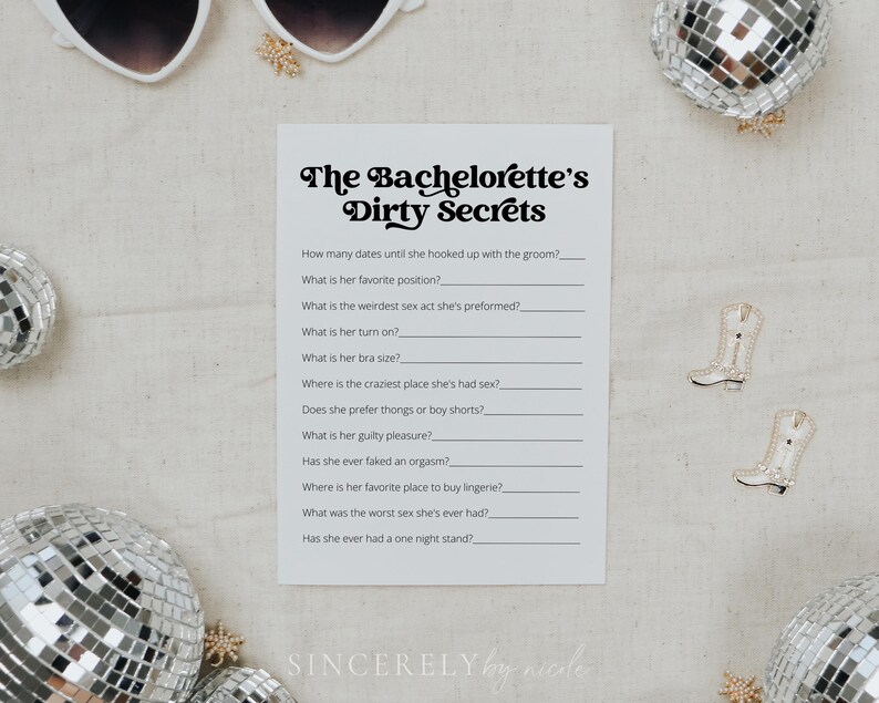 A printed game card for "The Bachelorette’s Dirty Secrets" resting on soft beige fabric with dried floral elements. A fun party activity designed for playful and candid moments.