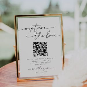 May include: A white sign in a gold frame with the text "capture the love" and a QR code. The sign also includes the text "SHARE YOUR PHOTOS WITH US. Please scan the QR code above and upload your photos!" and the names "samantha + jason" and the date "MAY 13.20XX".