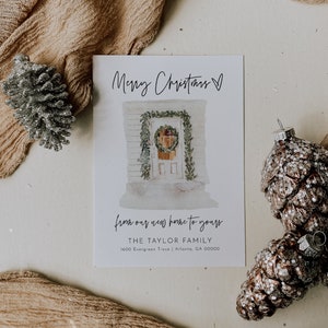 May include: A white Christmas card with a watercolor illustration of a house with a wreath on the door. The text "Merry Christmas" is written in black cursive at the top of the card. The text "from our new home to yours" is written in black cursive below the illustration. The text "THE TAYLOR FAMILY" is written in black below the cursive text. The text "1600 Evergreen Trace | Atlanta, GA 00000" is written in black below the family name. The text "SINCERELY BY NICOFF" is written in black at the bottom of the card.