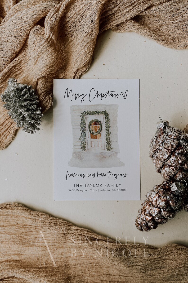 May include: A white Christmas card with a watercolor illustration of a house with a wreath on the door. The text "Merry Christmas" is written in black cursive at the top of the card. The text "from our new home to yours" is written in black cursive below the illustration. The text "THE TAYLOR FAMILY" is written in black below the cursive text. The text "1600 Evergreen Trace | Atlanta, GA 00000" is written in black below the family name. The text "SINCERELY BY NICOFF" is written in black at the bottom of the card.