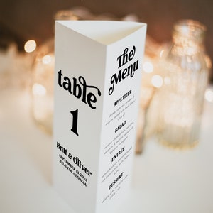 Op de afbeelding: Een witte tafelnummerkaart met zwarte tekst. De kaart is gevouwen in de vorm van een driehoek. De tekst op de kaart luidt "tafel 1" en "Britt & Oliver, 13 september 20xx, Atlanta, Georgia". De kaart bevat ook een menu met de kop "The Menu" en secties voor voorgerecht, salade, hoofdgerecht en dessert.