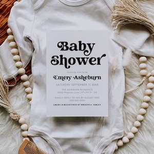 Op de afbeelding: Een witte baby romper met een witte kaart erop. De kaart heeft zwarte tekst die luidt "Baby Shower ter ere van de aanstaande moeder Emery Asheburn Zaterdag 17 september 20XX The Asheburn Residence 0000 Magnolia Lane | Atlanta, GA Graag reageren naar Ava voor 23 augustus 000-000-0000 Emery is geregistreerd bij Amazon & Target"