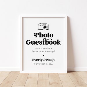 May include: A white framed sign with black text that reads "Photo Guestbook" with a camera icon above the text. The text below the title reads "snap a photo + leave us a message!" and "Everly & Noah" with the date "NOVEMBER 11, 20xx" below the names.