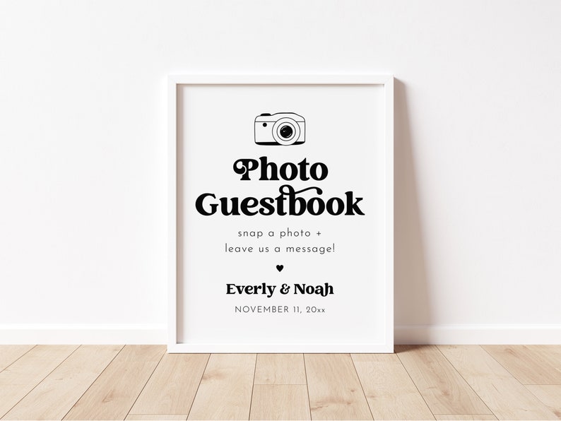 May include: A white framed sign with black text that reads "Photo Guestbook" with a camera icon above the text. The text below the title reads "snap a photo + leave us a message!" and "Everly & Noah" with the date "NOVEMBER 11, 20xx" below the names.