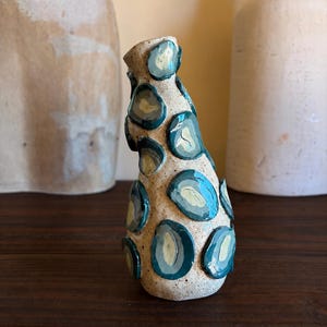 Puede incluir: Un pequeño jarrón de cerámica formado a mano con una base de color beige claro moteado. La superficie está decorada con parches circulares irregulares en relieve en tonos azul verdoso profundo y azul pálido, creando un aspecto texturizado.