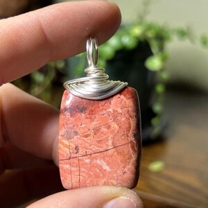 Può includere: Un ciondolo rettangolare con una pietra rossa e marrone, avvolto in filo d'argento. La pietra ha un aspetto naturale e strutturato con venature scure. Il filo è avvolto strettamente attorno alla parte superiore, formando un anello per una catena.