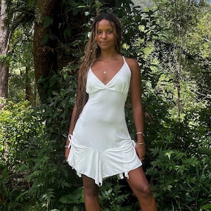 May include: A white halter dress with a ruffled hem is worn with black boots. The dress has a fitted bodice and a flowing skirt. The person is standing in a forest setting with tall trees and green foliage.