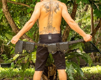 May include: Black leather belt with multiple pouches and gold-colored studs. The belt is held by a person wearing black shorts, standing in a lush, green outdoor setting. The belt has a unique, stylish design.