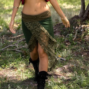 Puede incluir: Una mujer con un chaleco rojo con volantes sobre un top negro y una falda estampada en verde y marrón. Está de pie en una zona arbolada.