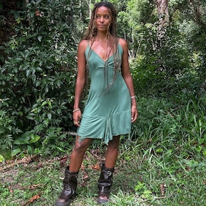 Puede incluir: Una mujer con un vestido verde salvia con tirantes finos y una falda drapeada está en un bosque. Lleva botas de cuero marrón oscuro. El fondo presenta un exuberante follaje verde y árboles.