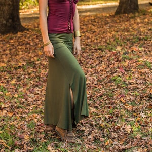 Puede incluir: Una mujer con un top burdeos con escote con cordones y mangas cortas, combinado con una falda larga verde oliva con abertura lateral. Lleva pulseras doradas. El fondo es un parque con árboles y hojas de otoño.