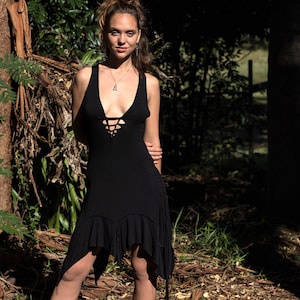 May include: A woman wearing a black, sleeveless dress with a lace-up detail at the front. The dress has a ruffled hem and is asymmetrical. She is standing in a wooded area and is wearing black boots.