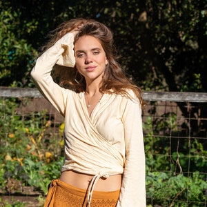 Puede incluir: Una mujer con un top de color crema con mangas largas y una falda de color mostaza con un patrón geométrico. Está de pie en un jardín con plantas verdes y una valla de madera en el fondo.