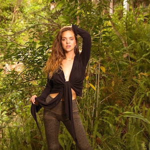 May include: A black tie-front top and olive green patterned bell-bottom pants are featured in this image. The pants have a floral design and flared bottoms. The model is standing in a lush, green forest setting.