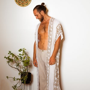 Puede incluir: Una t&uacute;nica blanca de manga larga con un patr&oacute;n geom&eacute;trico marr&oacute;n. La t&uacute;nica est&aacute; abierta por delante y tiene un dise&ntilde;o largo y fluido. La persona que lleva la t&uacute;nica est&aacute; de pie sobre una alfombra estampada.