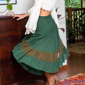 Puede incluir: Una mujer con un top blanco de hombros descubiertos y una falda verde de capas con ribete de encaje. La falda es de estilo envolvente y tiene una silueta larga y fluida.