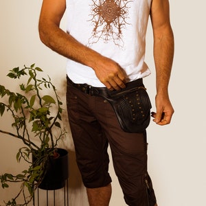 K&ouml;nnte beinhalten: Eine Person tr&auml;gt ein wei&szlig;es &auml;rmelloses Shirt mit einem braunen Grafikdesign und braune Shorts. Sie tr&auml;gt eine schwarze Leder-Bauchtasche mit Metallnieten.