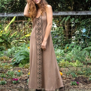 May include: A woman wearing a brown maxi dress with a black geometric pattern. She is standing in front of a wooden fence with a wire mesh background. The dress has a relaxed fit and a flowy silhouette.