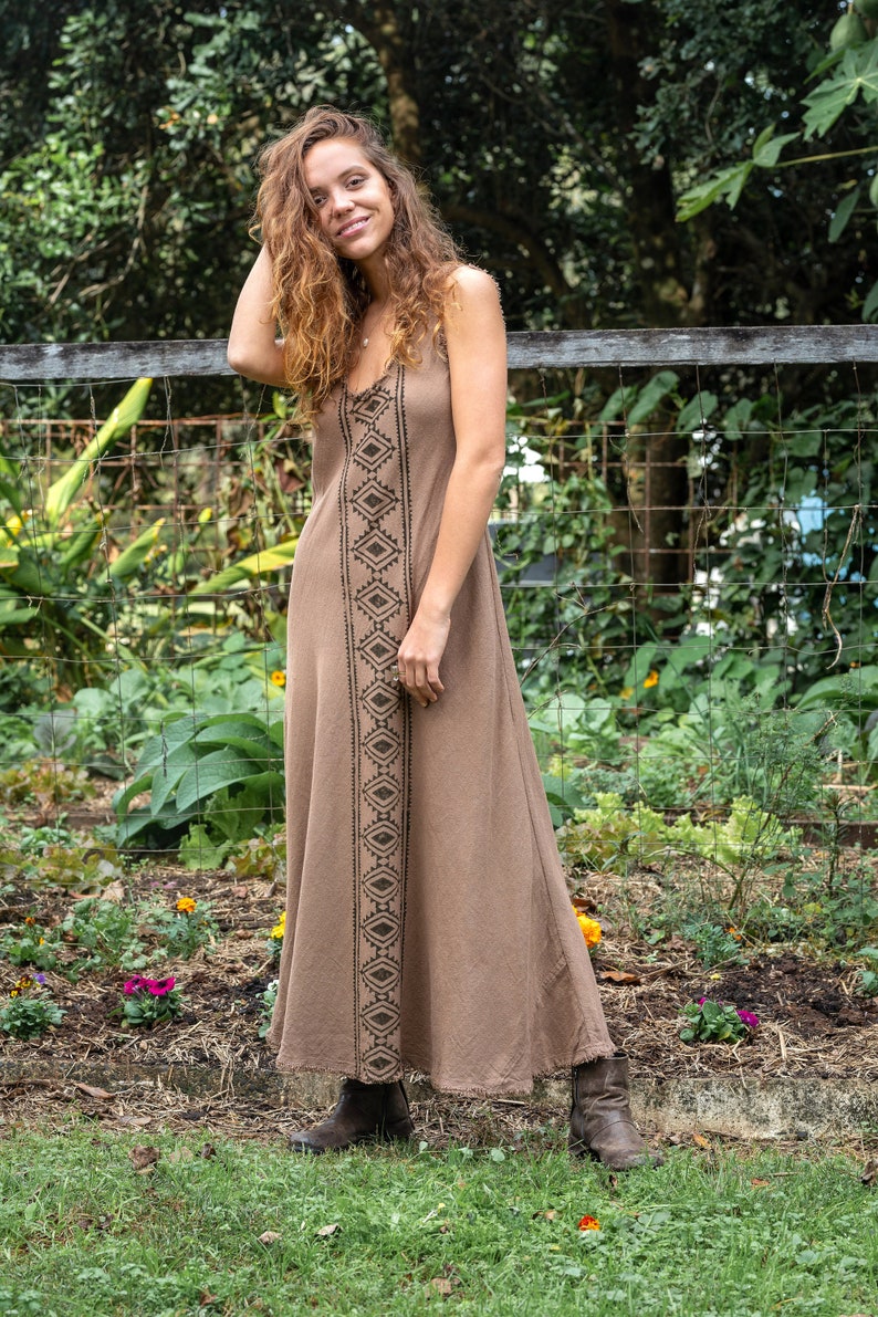 May include: A woman wearing a brown maxi dress with a black geometric pattern. She is standing in front of a wooden fence with a wire mesh background. The dress has a relaxed fit and a flowy silhouette.