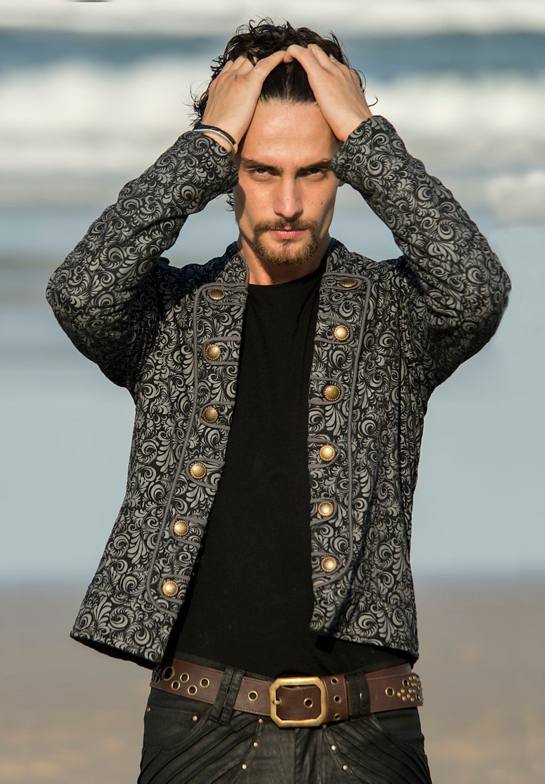 Puede incluir: Un hombre con una chaqueta gris estampada con botones dorados, una camiseta negra y pantalones vaqueros negros con un cintur&oacute;n marr&oacute;n y detalles dorados.