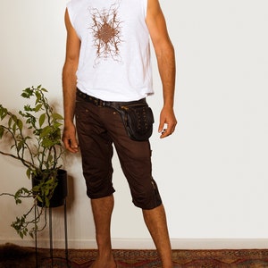 K&ouml;nnte beinhalten: Ein Mann tr&auml;gt ein wei&szlig;es &auml;rmelloses Hemd mit einem braunen geometrischen Muster, braune Cargo-Shorts und einen schwarzen Lederg&uuml;rtel mit einer Nietentasche. Er steht auf einem gemusterten Teppich.