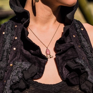 May include: A gold-toned necklace featuring a teardrop-shaped pink stone pendant suspended from a thin chain, paired with ornate gold-toned filigree hoop earrings. The jewellery is worn with a black sleeveless vest with ruffled edges and metal studs.