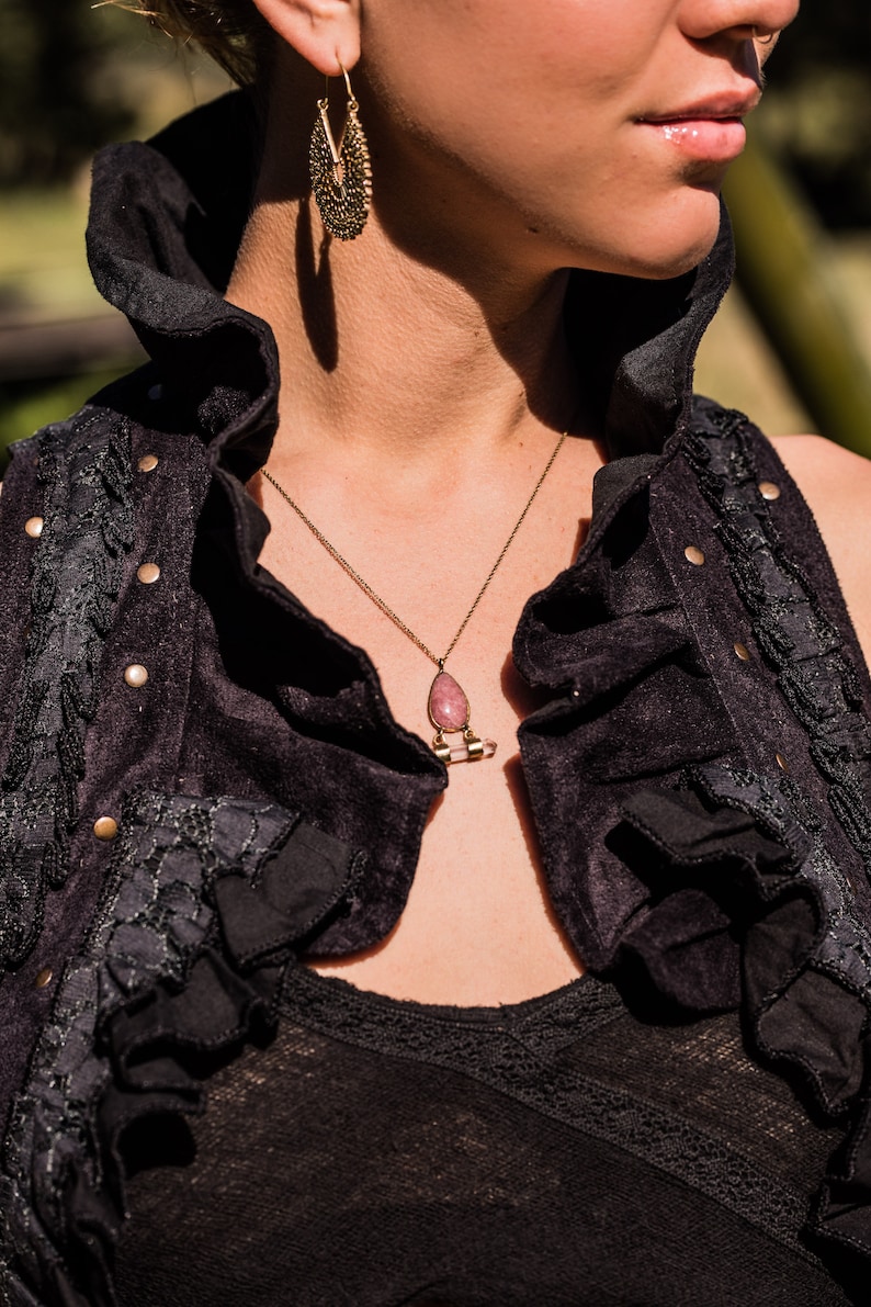 May include: A gold-toned necklace featuring a teardrop-shaped pink stone pendant suspended from a thin chain, paired with ornate gold-toned filigree hoop earrings. The jewellery is worn with a black sleeveless vest with ruffled edges and metal studs.