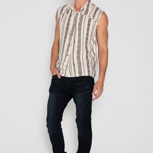 May include: A sleeveless button-up shirt with a white background and black vertical stripes. The shirt has a geometric pattern and a collar. The model is wearing dark blue jeans and brown boots.