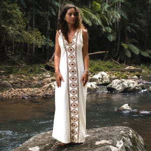 May include: A white maxi dress with a brown geometric pattern down the front. The dress is sleeveless and has a V-neckline. The model is standing in a natural setting with a river and rocks in the background.