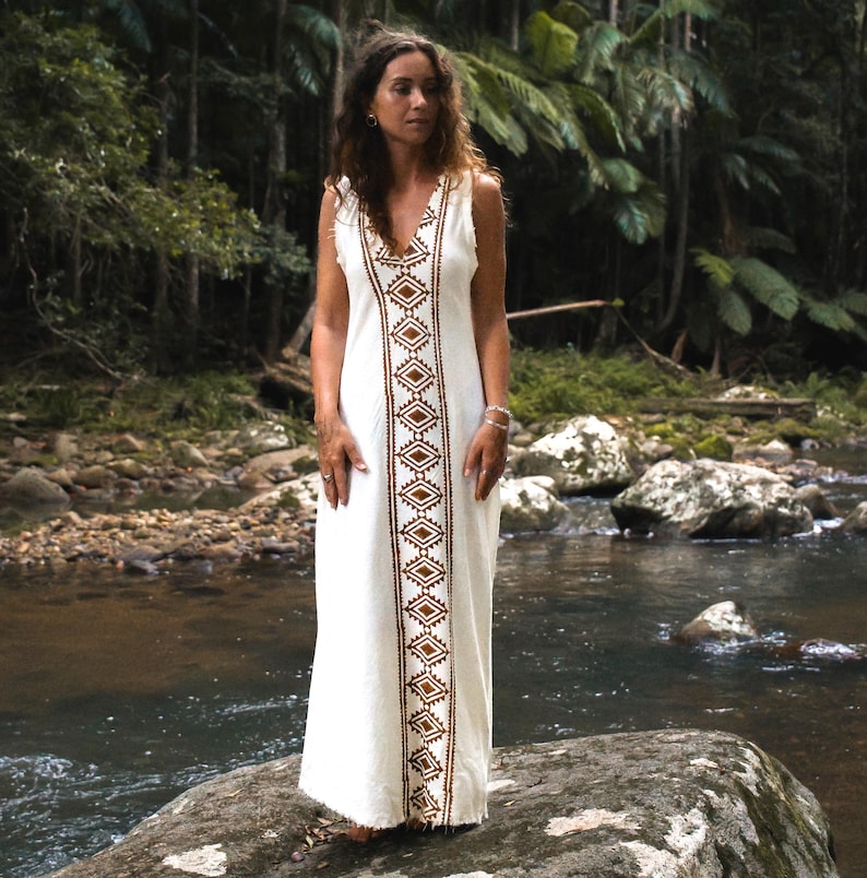 May include: A white maxi dress with a brown geometric pattern down the front. The dress is sleeveless and has a V-neckline. The model is standing in a natural setting with a river and rocks in the background.