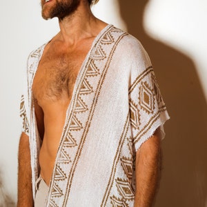 Puede incluir: Un poncho blanco de crochet con un patr&oacute;n geom&eacute;trico marr&oacute;n. El poncho tiene cuello en V y mangas cortas.