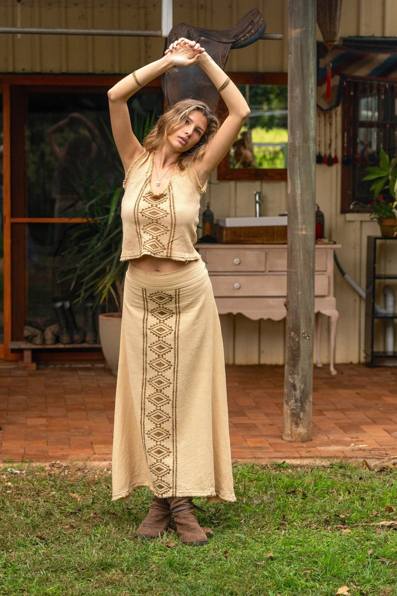 May include: A woman wearing a beige top and skirt with brown geometric embroidery. The skirt is long and flowy, and the top is cropped. She is standing in a grassy area with a wooden structure in the background.