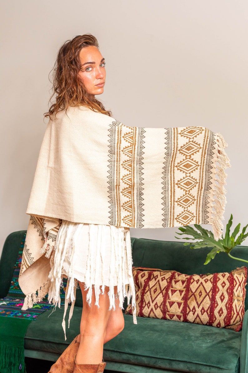 May include: A woman wearing a white and brown patterned poncho with fringe. She is standing in front of a green couch with a red and brown patterned pillow.