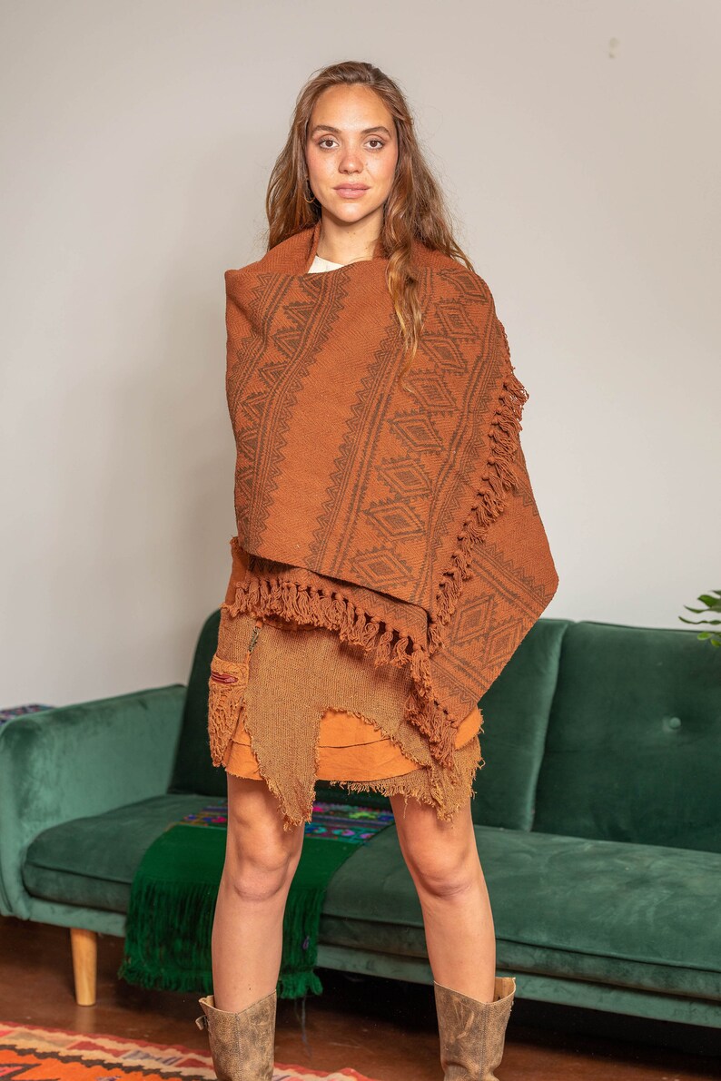 May include: A woman wearing a brown poncho with a geometric pattern and fringe. The poncho is draped over a brown skirt with frayed edges. She is standing in front of a green couch.