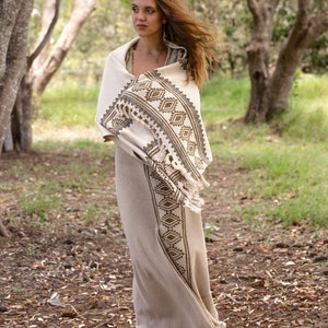 May include: A woman wearing a long, white dress with a brown and white geometric pattern. She is standing in a forest with trees and green grass in the background.