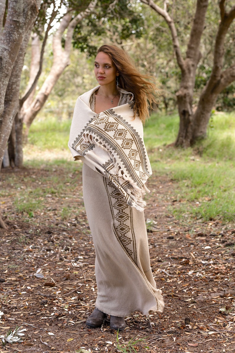 May include: A woman wearing a long, white dress with a brown and white geometric pattern. She is standing in a forest with trees and green grass in the background.