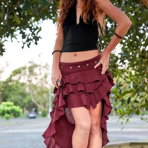 May include: A woman wearing a black halter top and a burgundy ruffled skirt with a high-low hemline. She is standing in front of a tree and has her hand on her hip.