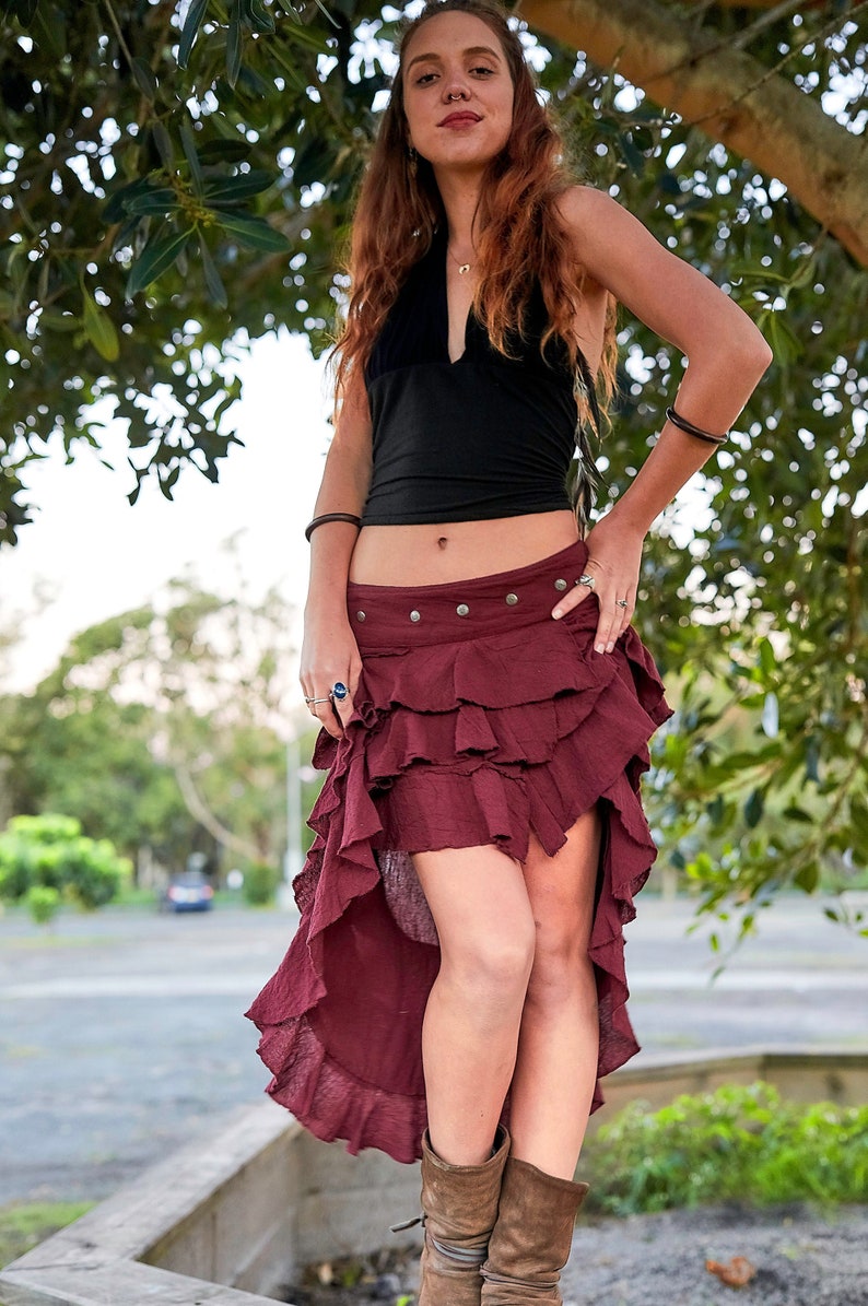 May include: A woman wearing a black halter top and a burgundy ruffled skirt with a high-low hemline. She is standing in front of a tree and has her hand on her hip.