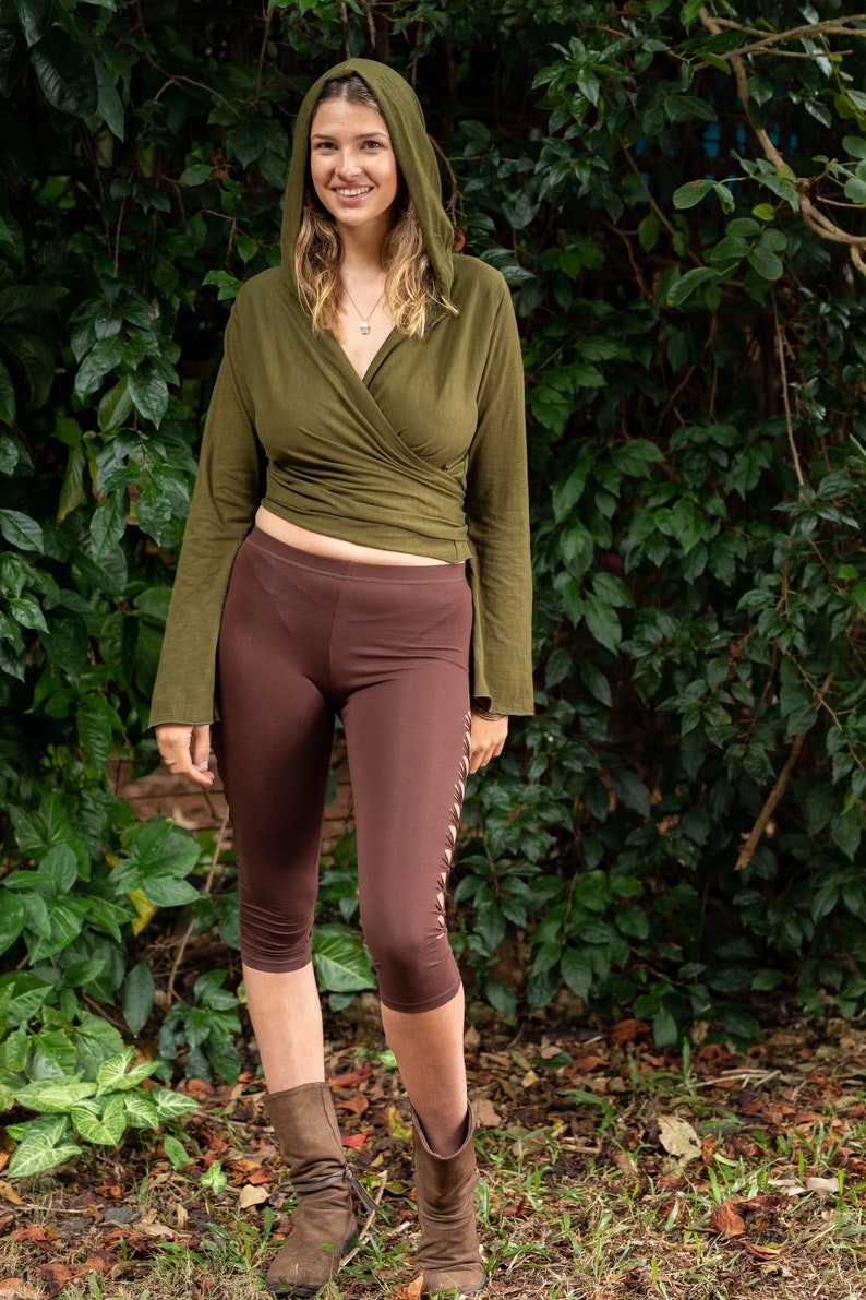 May include: A woman wearing a green hooded wrap top with long sleeves and brown leggings. She is standing in front of a green leafy background.