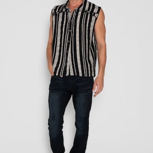 May include: A sleeveless button-up shirt with black and white vertical stripes. The shirt has a collar and a button closure. The model is wearing dark blue jeans and brown shoes.