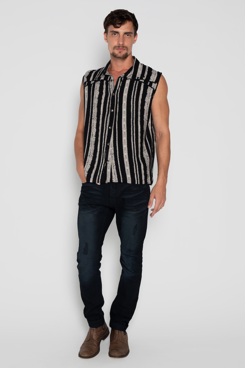 May include: A sleeveless button-up shirt with black and white vertical stripes. The shirt has a collar and a button closure. The model is wearing dark blue jeans and brown shoes.