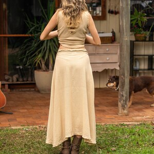 May include: A woman wearing a light brown, sleeveless top and a long, flowing skirt. The skirt is made of a lightweight fabric and has a frayed hem. She is wearing brown boots and standing on a grassy lawn.