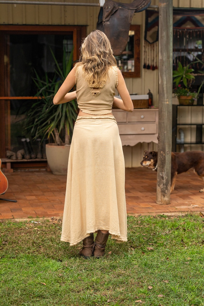 May include: A woman wearing a light brown, sleeveless top and a long, flowing skirt. The skirt is made of a lightweight fabric and has a frayed hem. She is wearing brown boots and standing on a grassy lawn.