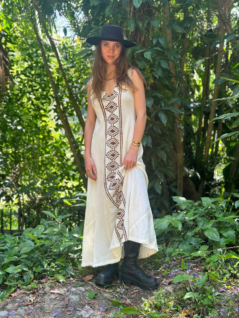 May include: A white maxi dress with brown geometric embroidery. The dress has a V-neckline and a sleeveless design. The model is wearing a black hat and black boots.