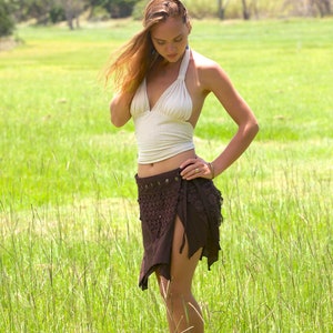 May include: A woman wearing a white halter top and a brown crochet skirt with a beaded belt. She is standing in a field of tall grass.