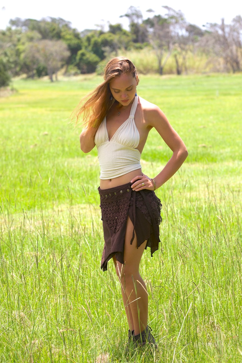 May include: A woman wearing a white halter top and a brown crochet skirt with a beaded belt. She is standing in a field of tall grass.